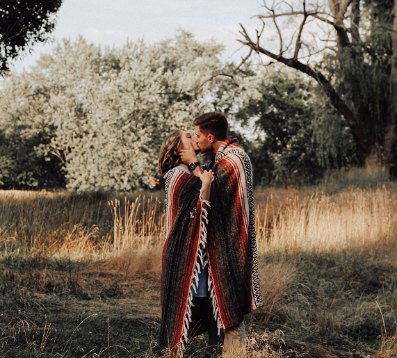 couple standing in a forest who has a blanket wrapped around them, kissing each other, symbolizing what does it mean to be vulnerable in a relationship