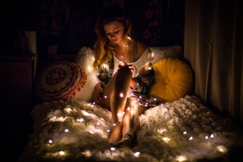 woman in t-shirt, sitting in the darkness on her bed with hundreds of little lights on, vibrational energy of love