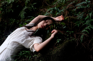 woman with dark hair and white dress, lying on a bed of ferns, divine feminine energy