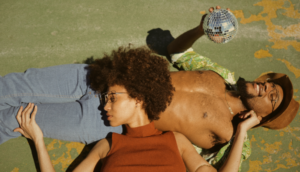 An African American couple laying on the concrete floor, her head on his lap, he has a disco ball in his hand